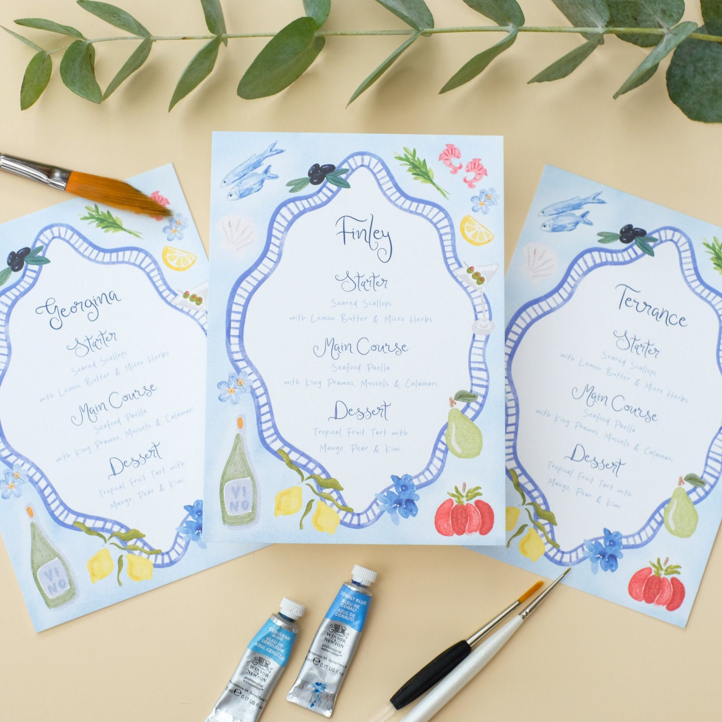 Hand painted wedding menu with blue border and watercolour illustrations of lemons, fish, shrimps and olives